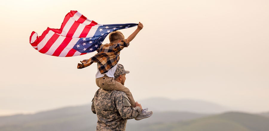 A Military member with a kid on his shoulder, while the kid is holding an american flag 